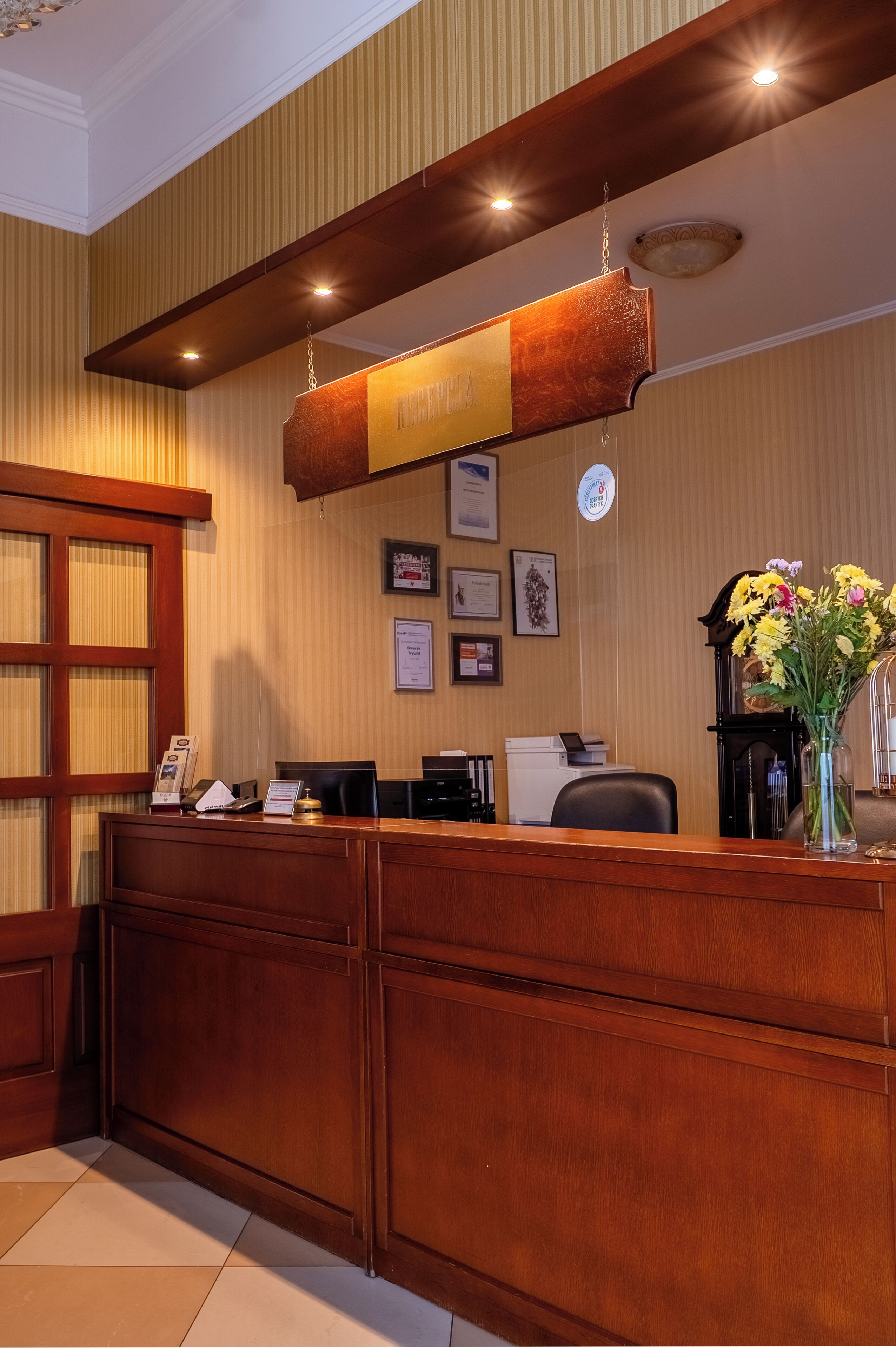 Un mostrador de recepción con detalles de madera, certificados enmarcados en la pared y un jarrón con flores.