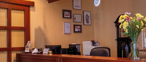 Un mostrador de recepción con detalles de madera, certificados enmarcados en la pared y un jarrón con flores.