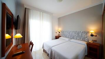 Habitació d'hotel amb dos llits, una taula de treball amb un comandament a distància, una cadira, un televisor i un capçal de llit amb un disseny decoratiu.