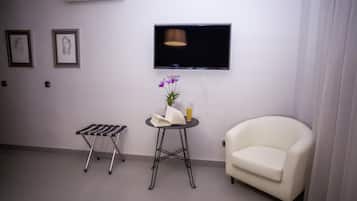 Una habitación con una televisión de pantalla plana montada en la pared, una mesita redonda con un libro y un jarrón de flores, un sillón blanco y una silla plegable.