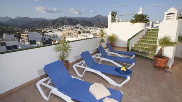 A rooftop terrace with blue lounge chairs, a staircase, and a view of a town and mountains. - Hotel Bajamar (Nerja)