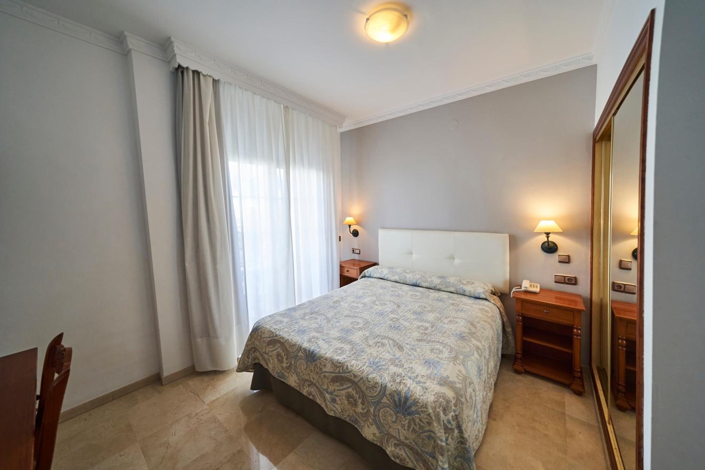 Un dormitorio con cama, mesitas de noche, espejo y ventana con cortinas.