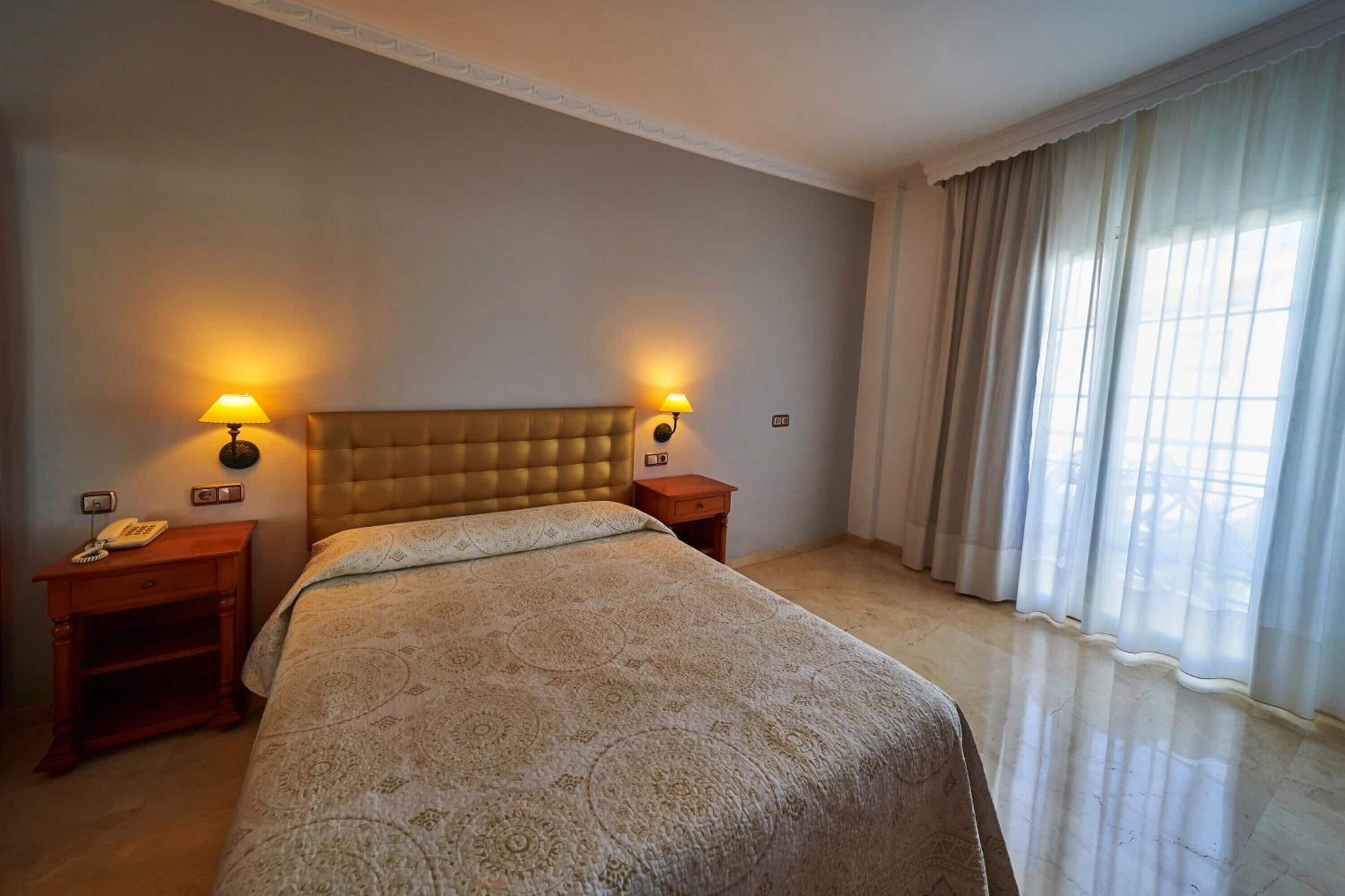 Une chambre d’hôtel avec un grand lit, deux tables de chevet, un téléphone et une fenêtre avec des rideaux.