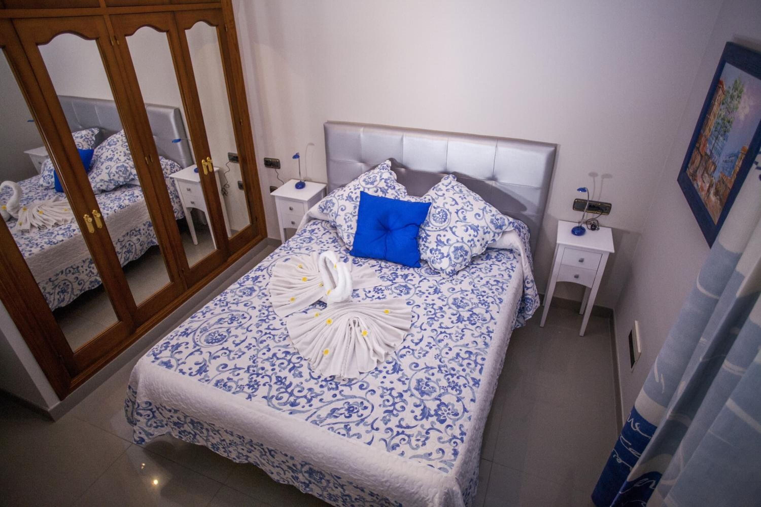 A neatly made bed with a blue and white patterned bedspread, a blue pillow, and a decorative swan towel. There is a wooden wardrobe with mirrored doors and a white bedside table with a lamp.
