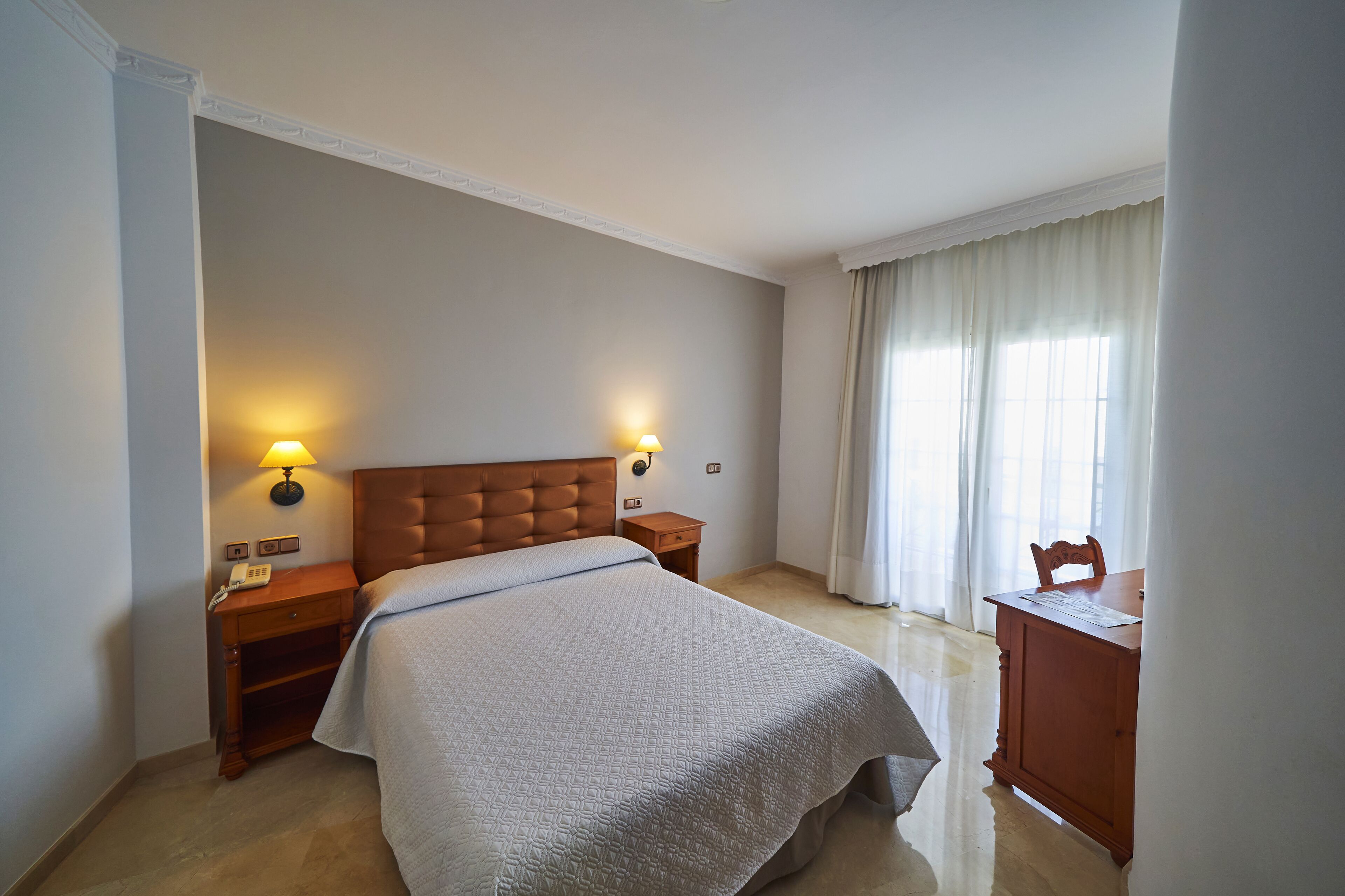 A hotel room with a large bed, two bedside lamps, a wooden nightstand, a wooden dresser, and a window with curtains.