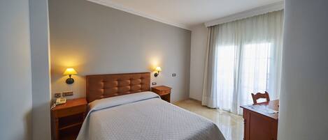 A hotel room with a large bed, two bedside lamps, a wooden nightstand, a wooden dresser, and a window with curtains.