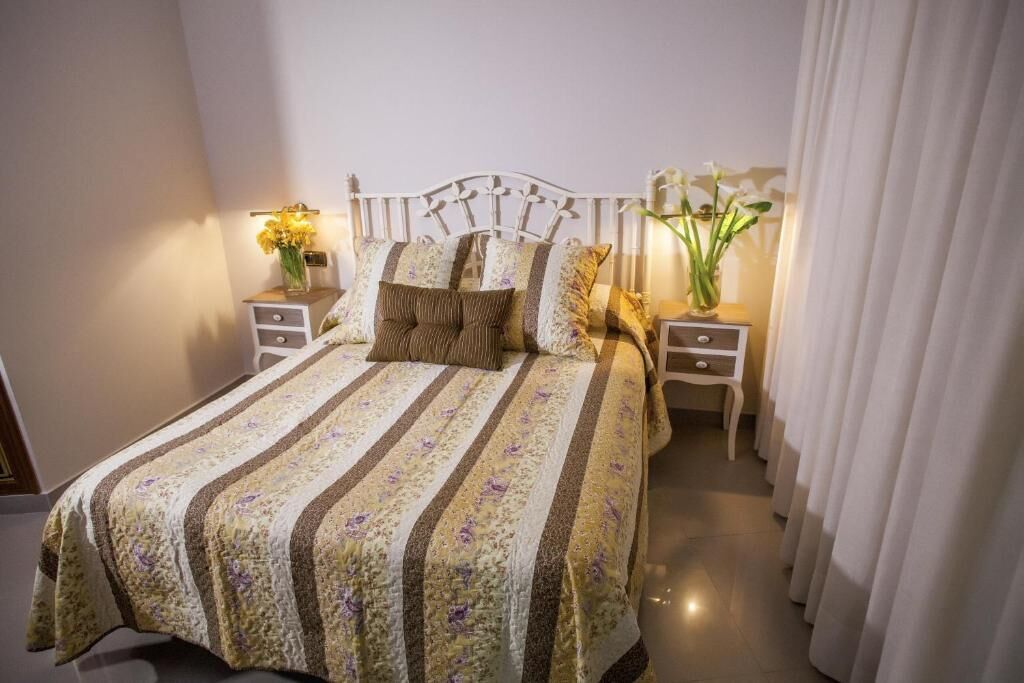Una cama bien tendida con una colcha estampada, dos mesitas de noche con lámparas y una cortina a la derecha.