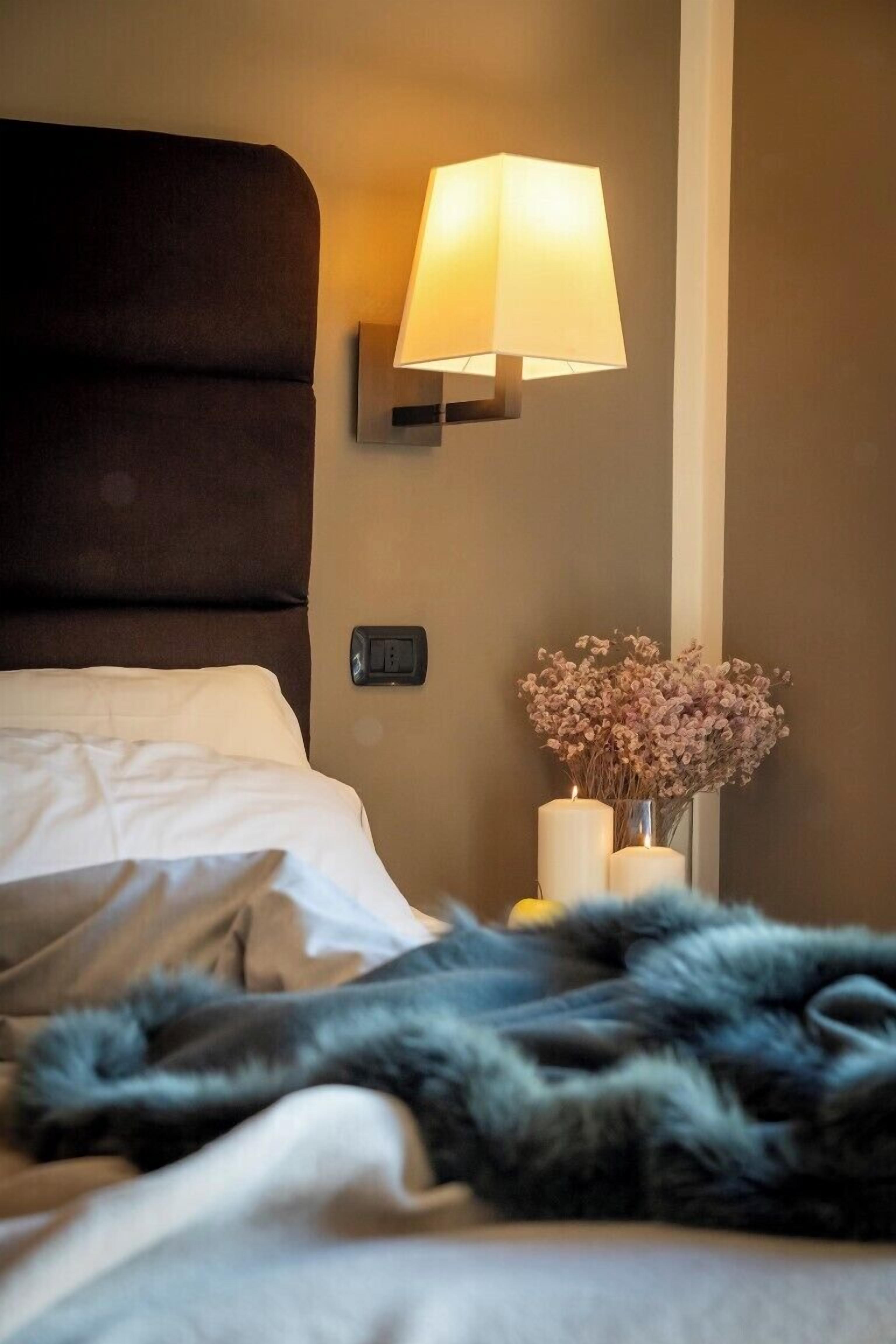 Una camera d'albergo con un letto, una lampada a parete, un vaso con fiori secchi e una presa elettrica.