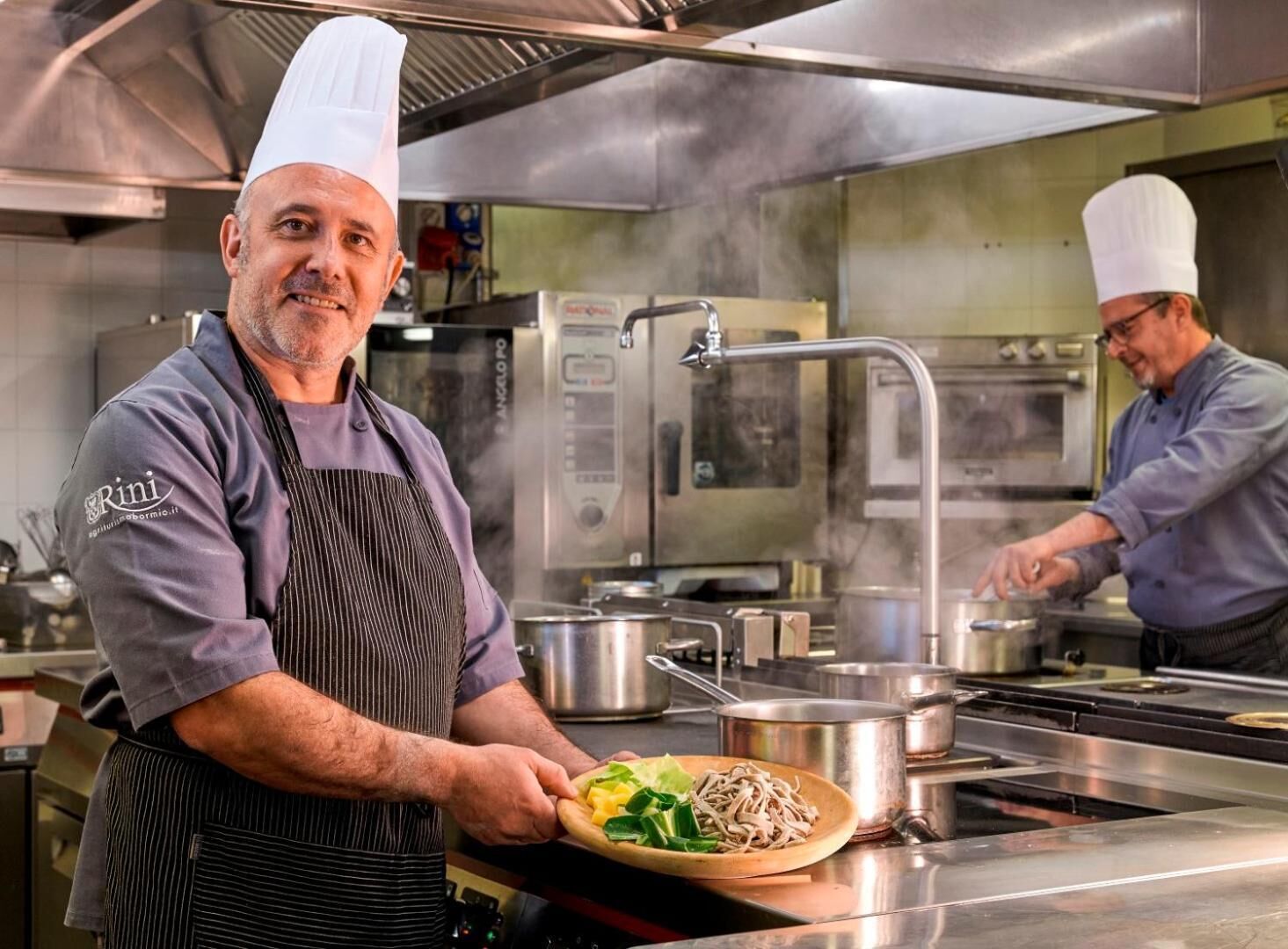 Un chef en una cocina con electrodomésticos de acero inoxidable, un horno grande y una olla sobre la encimera.