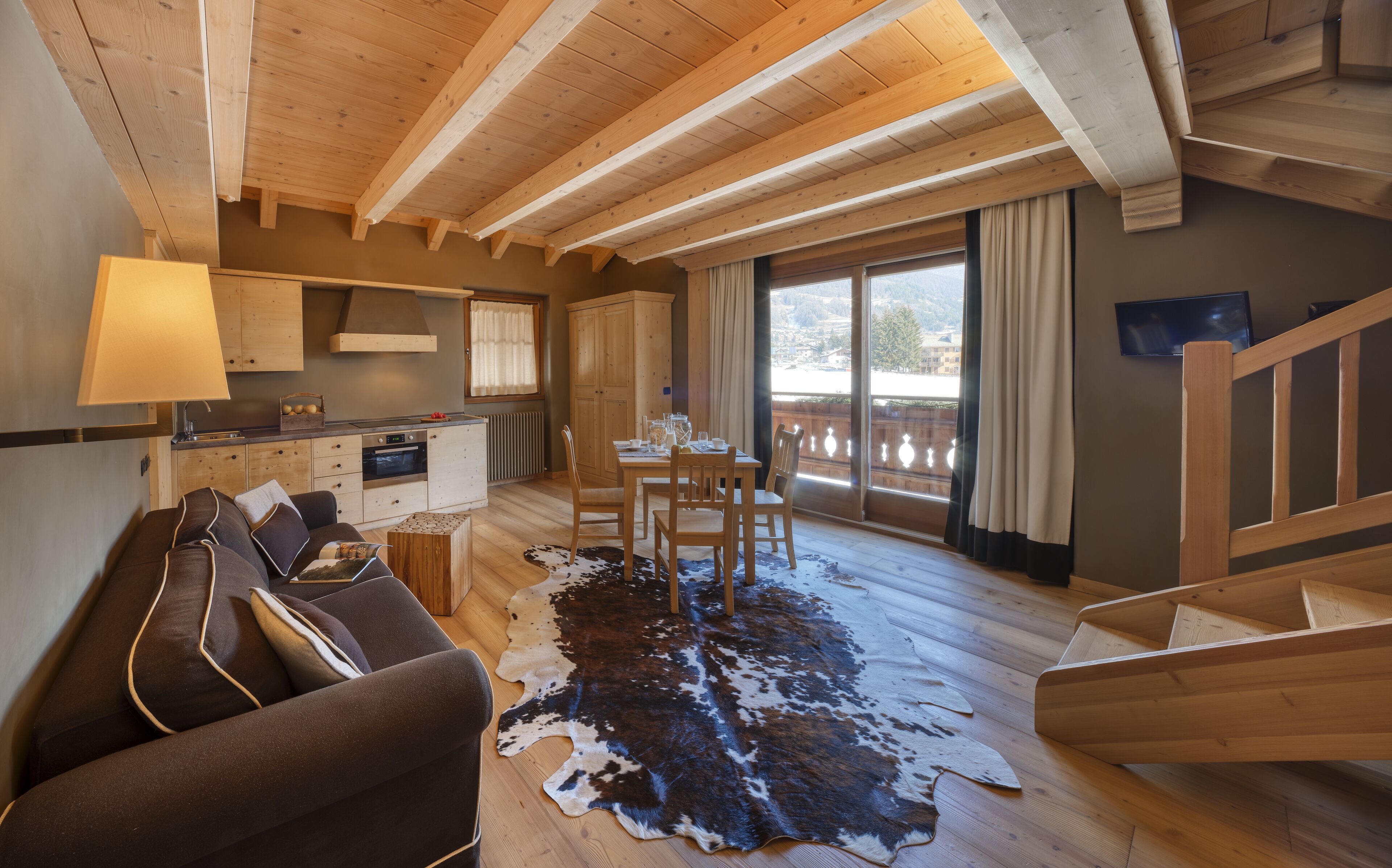A cozy cabin interior with a wooden ceiling, a dining area with a table and chairs, a kitchen with a stove and cabinets, and a large window with a view.
