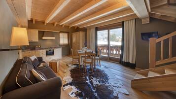 A cozy cabin interior with a wooden ceiling, a dining area with a table and chairs, a kitchen with a stove and cabinets, and a large window with a view.