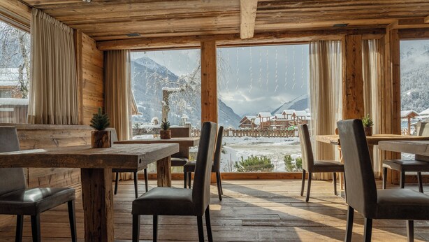 Interior de madeira com mesas e cadeiras, grandes janelas com cortinas e vista para montanhas nevadas.