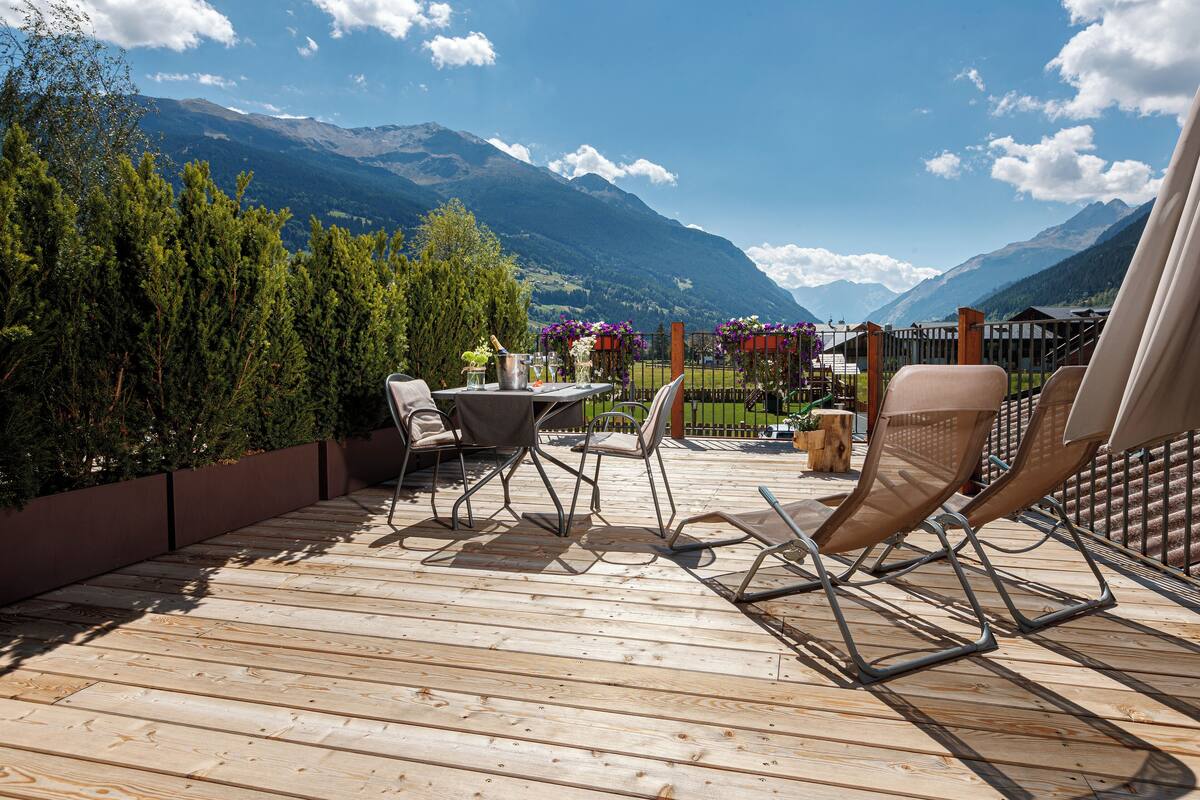 A wooden deck with outdoor furniture, a table, and chairs, overlooking a mountainous landscape.