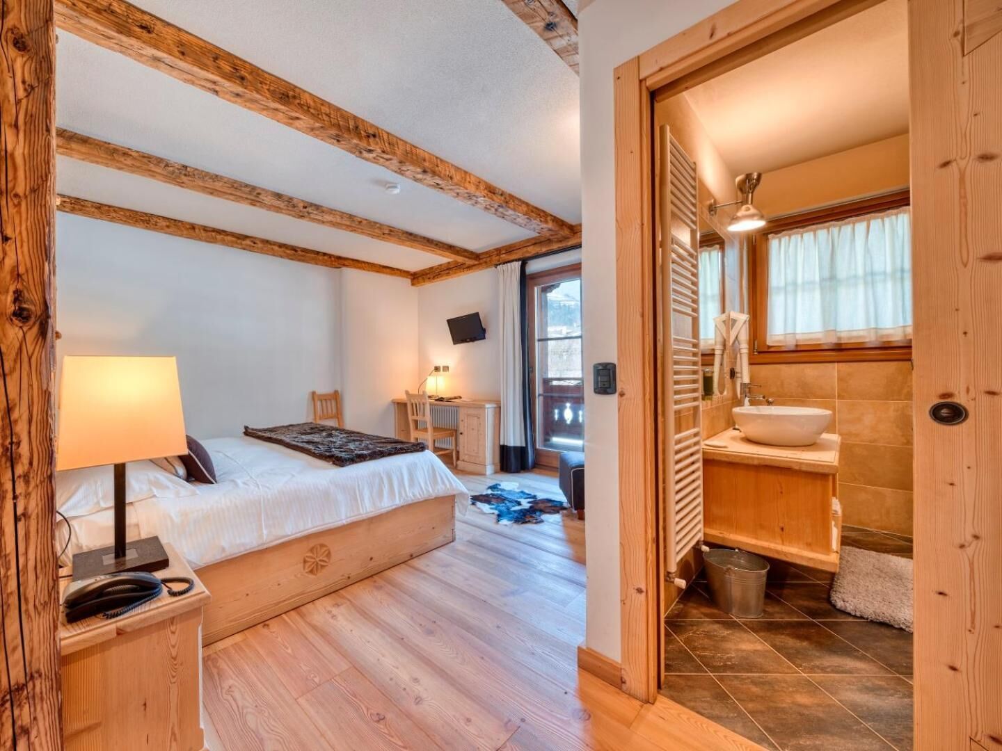 Un dormitorio con techo de madera, una cama, un escritorio, una silla, un televisor y un baño con lavabo y ventana.