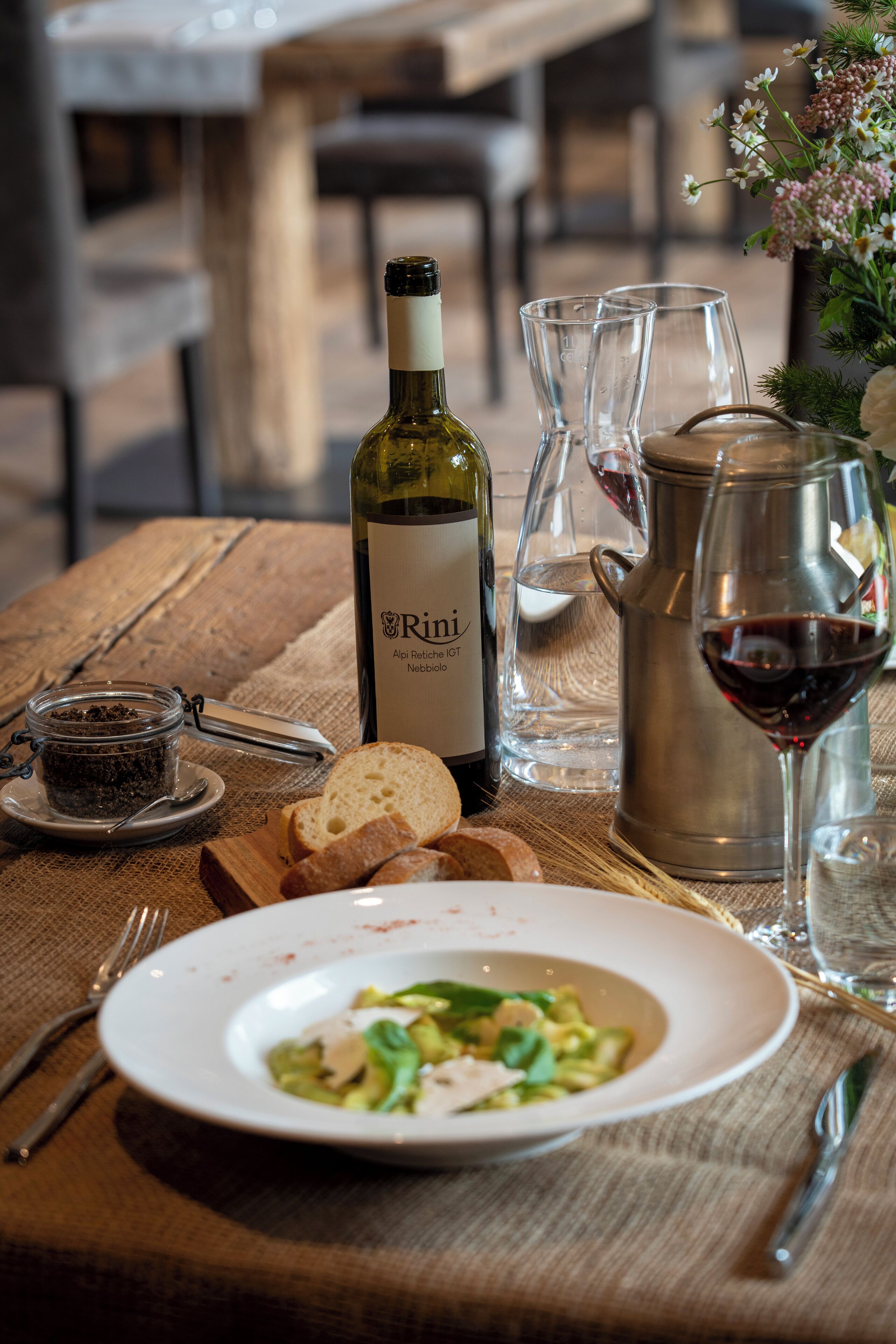 A wooden table with a bottle of wine, glasses, a pitcher, bread, and a plate of food.