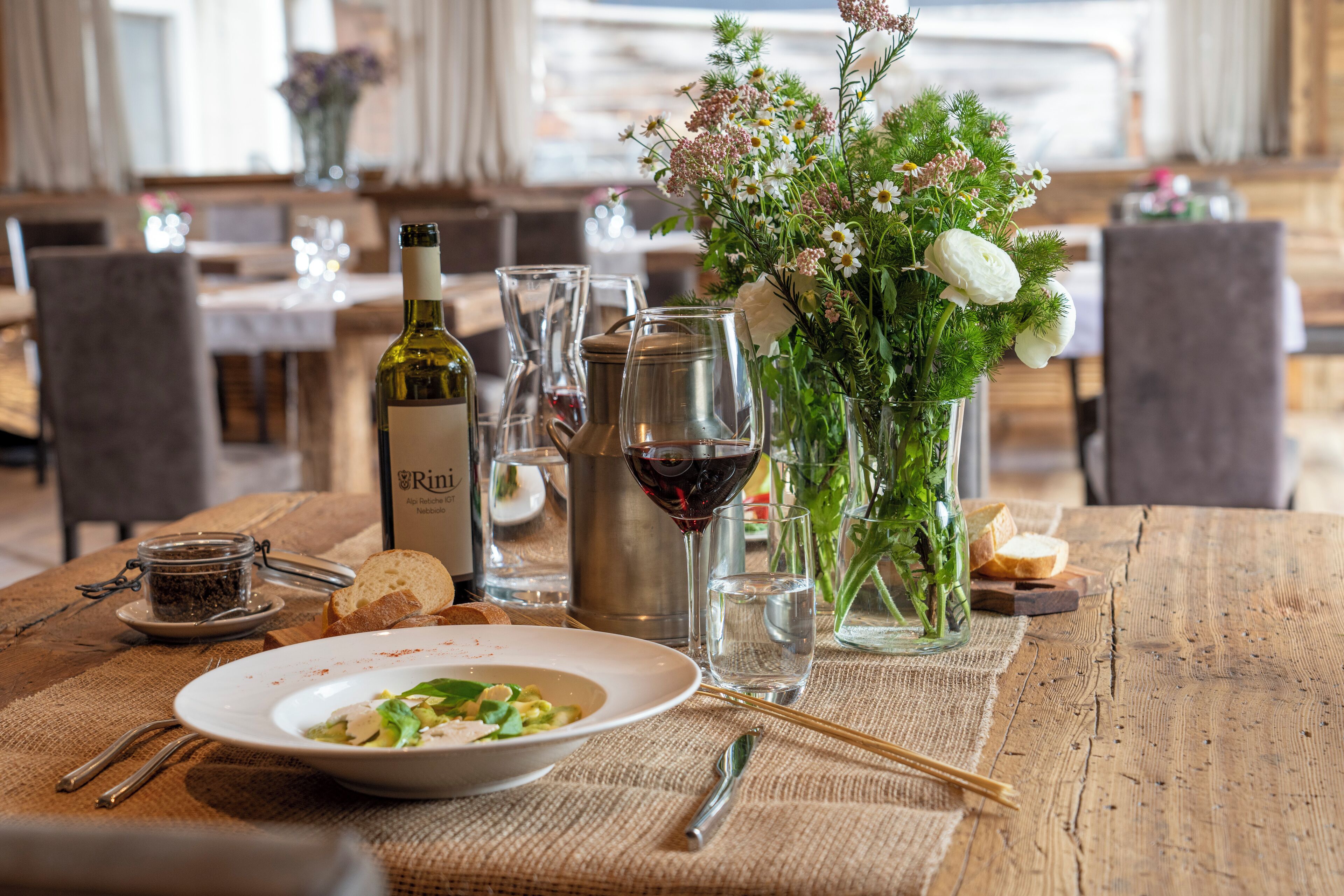 A dining table set with a bottle of wine, a glass, a vase with flowers, and a plate of food.