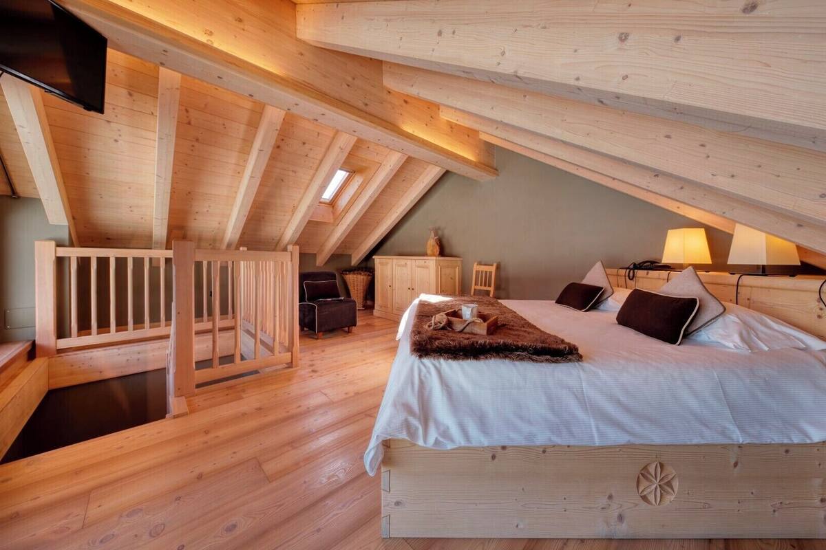A high-ceilinged attic bedroom with a wooden bed, a small seating area, and a window.