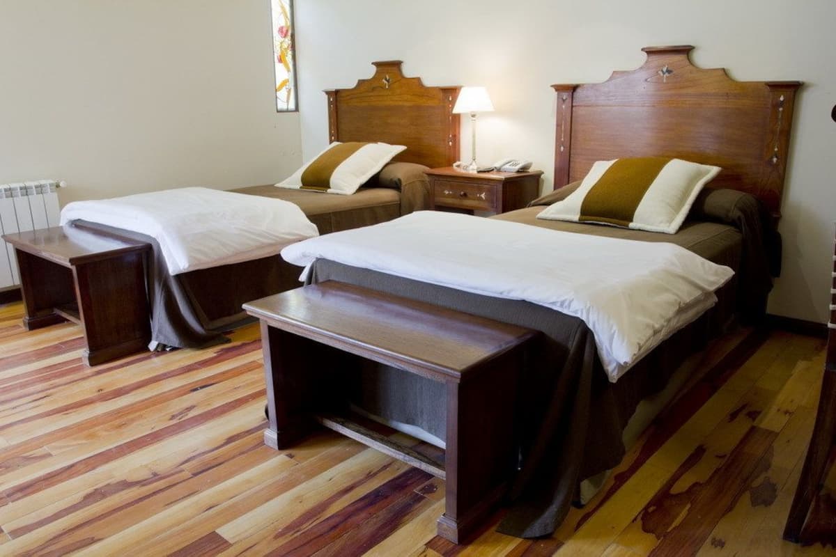 Habitación de hotel con dos camas individuales, cabeceros de madera, mesitas de noche y un banco.
