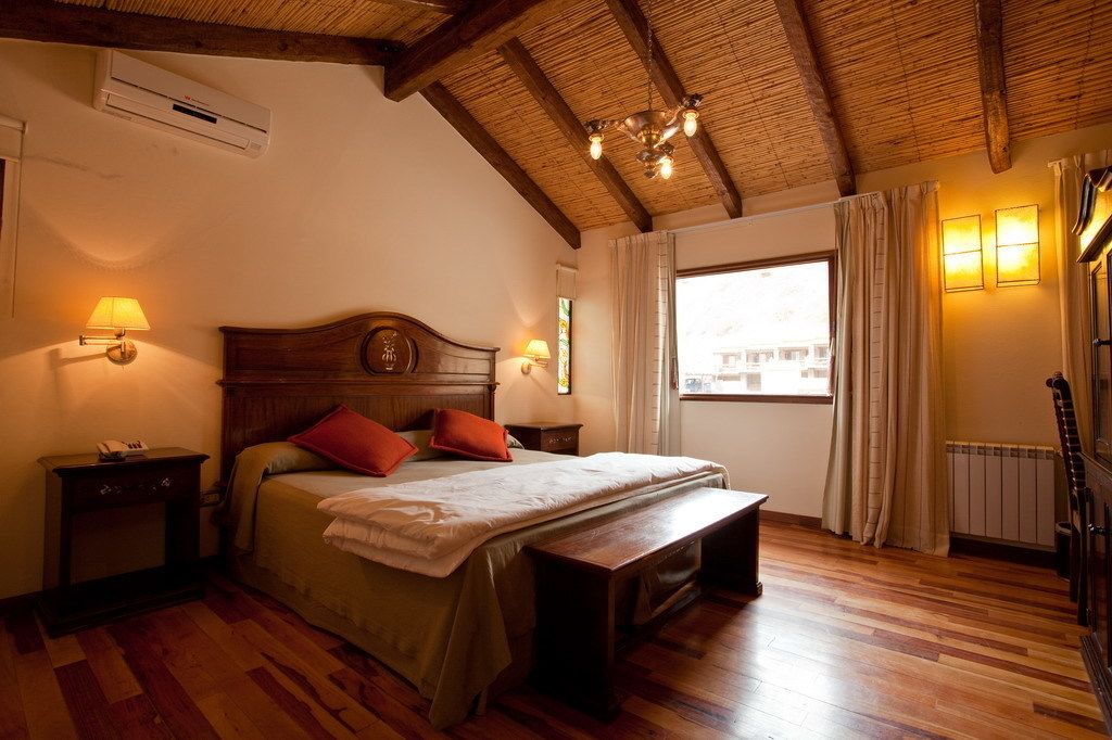 A bedroom with a wooden ceiling, a bed with pillows, a nightstand, a bench, and a window with curtains.