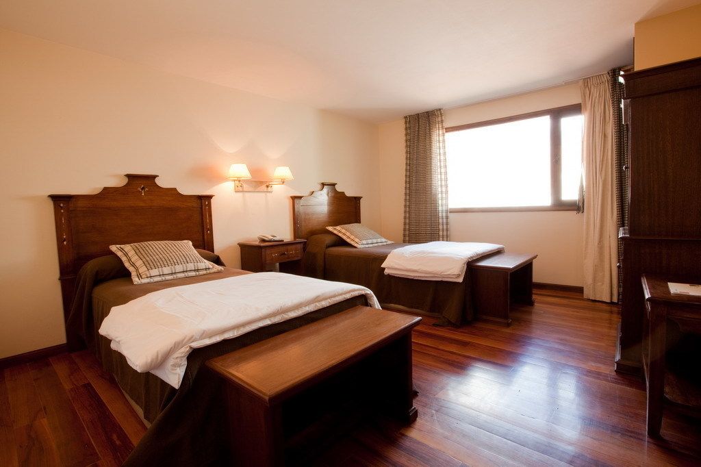 A hotel room with two beds, wooden headboards, bedside tables, and a window with curtains.