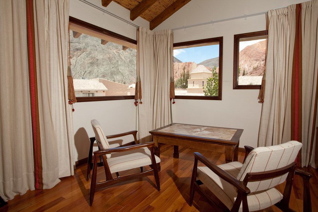 A room with a wooden floor, a table, and two chairs. There are large windows with curtains, and a view of mountains and buildings outside.