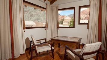 A room with a wooden floor, a table, and two chairs. There are large windows with curtains, and a view of mountains and buildings outside.