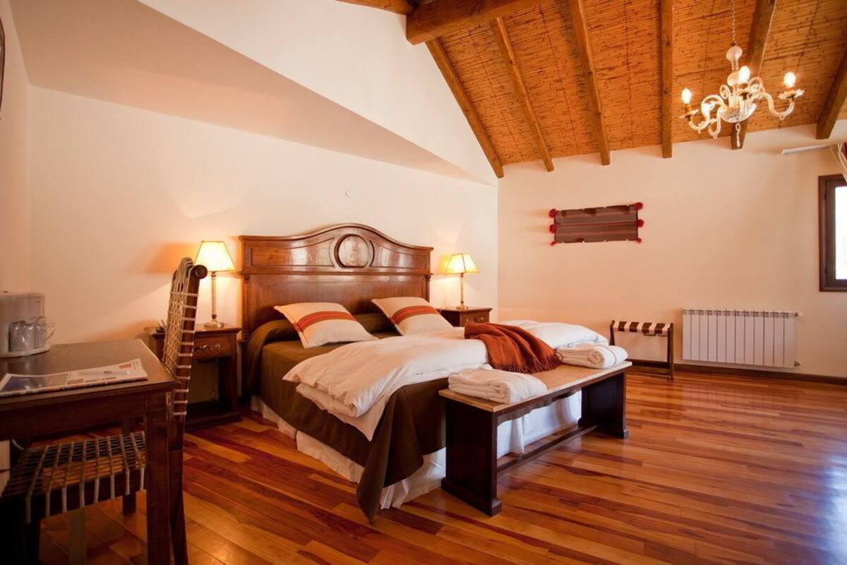 Un dormitorio con una cama de madera, dos mesitas de noche con lámparas, un banco de madera, un radiador y una araña.