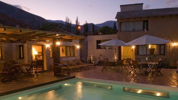 A pool area with lounge chairs and umbrellas, a wooden deck, and a building with multiple windows and doors. - Marques de Tojo Hotel (Purmamarca)