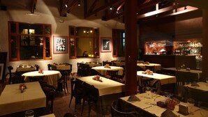 Restaurante com várias mesas e cadeiras, vigas de madeira no teto e área de bar visível através de um painel de vidro.