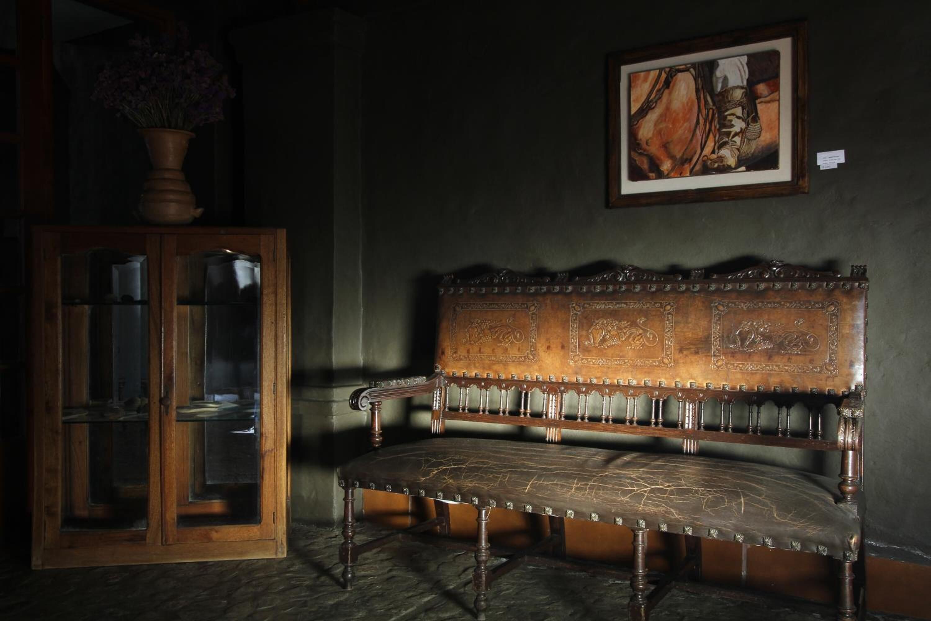 A wooden bench with intricate carvings, a glass cabinet with items inside, and a framed painting on the wall.