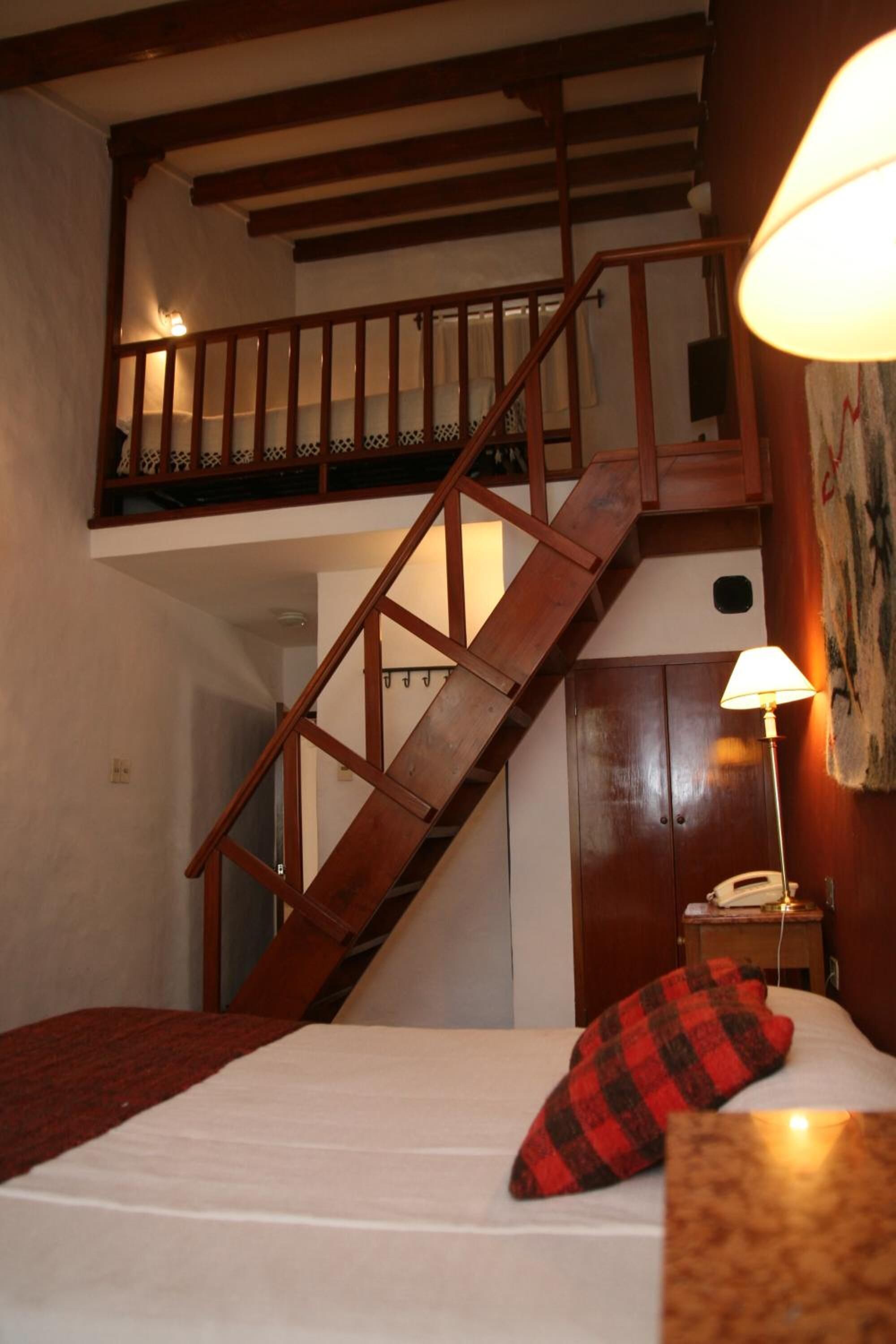 A bedroom with a bed, a red and black checkered pillow, a nightstand with a lamp, and a wooden staircase leading to an upper level.
