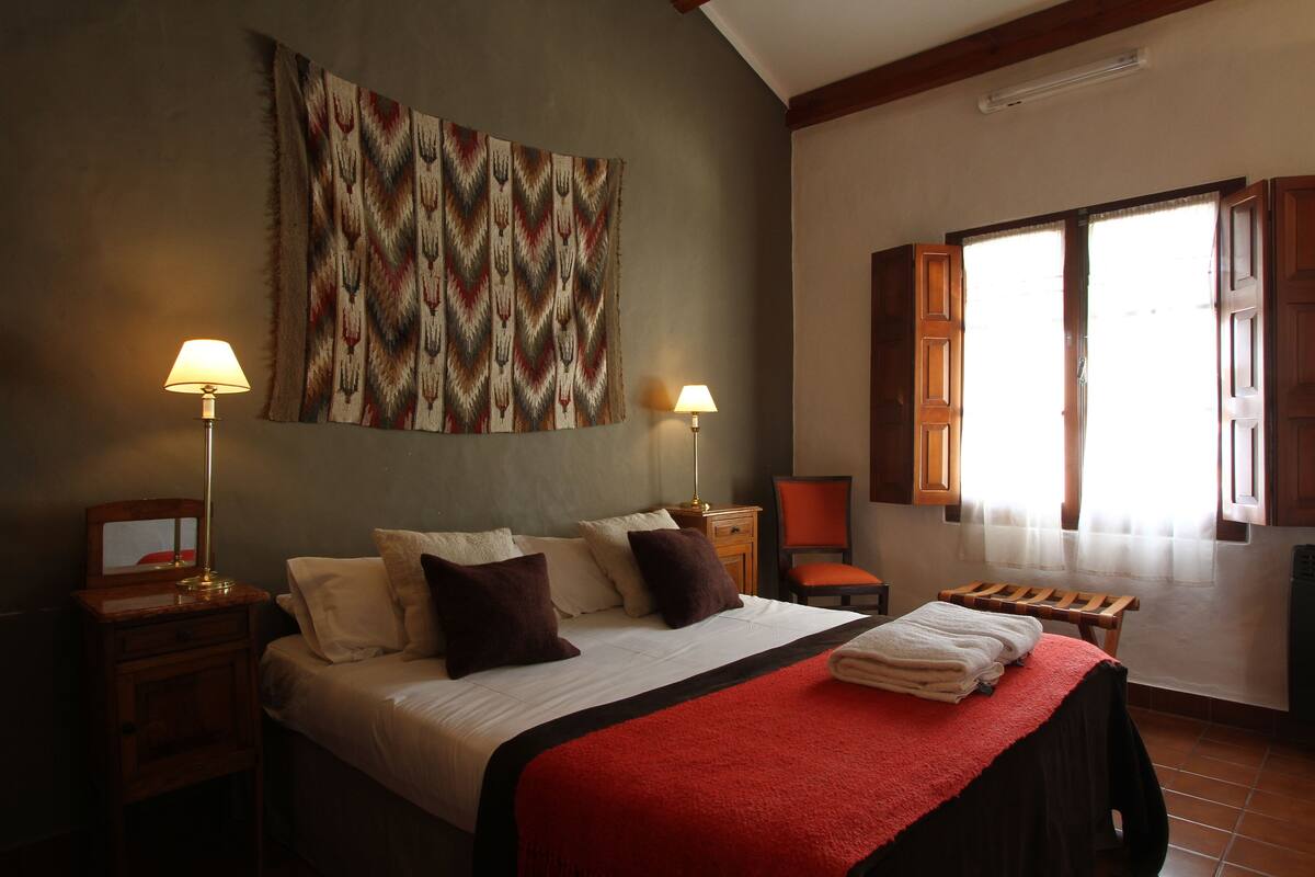 Une chambre d’hôtel avec un grand lit, deux lampes de chevet, un plaid rouge, un banc en bois et une grande fenêtre avec des rideaux.