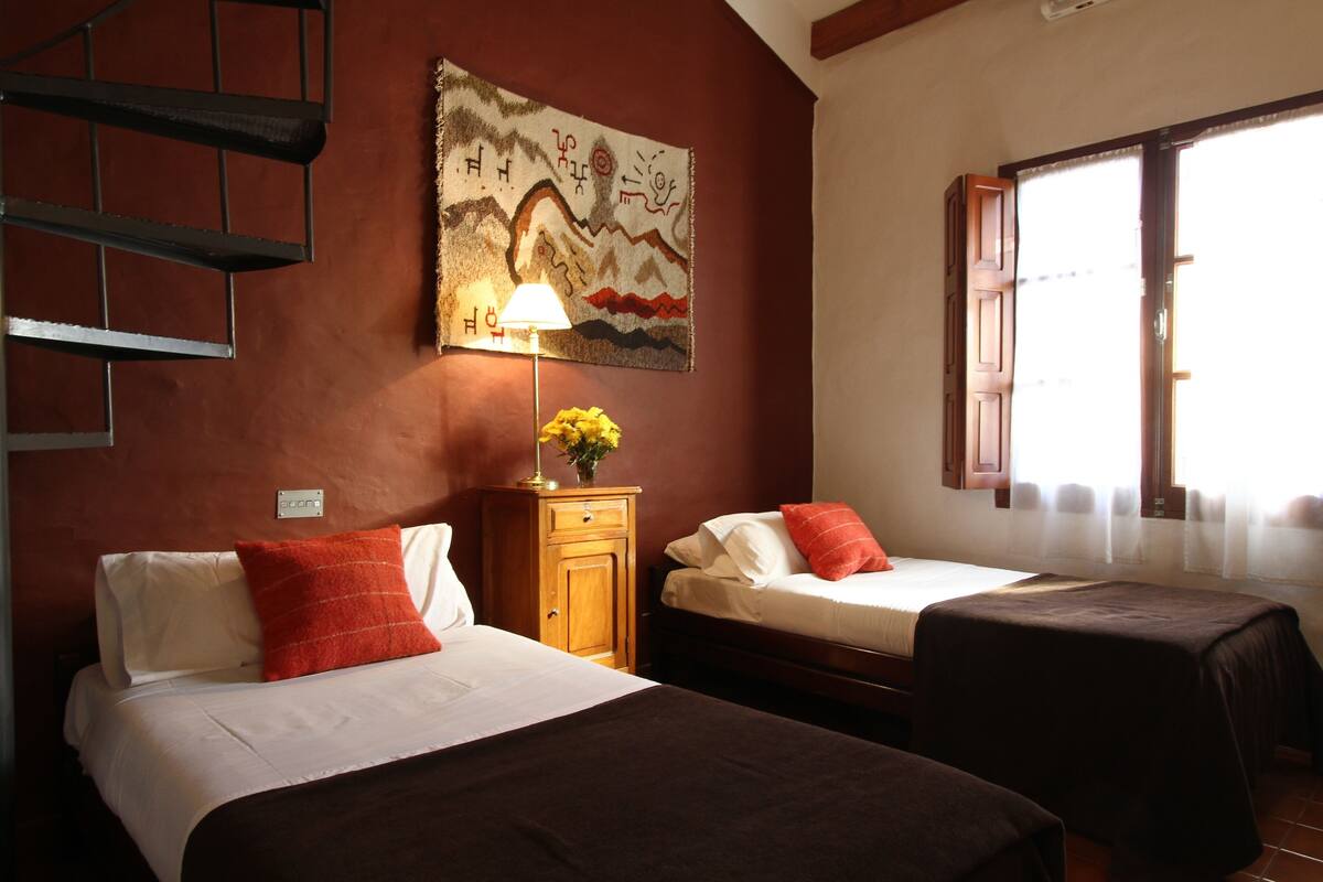 Une chambre d’hôtel avec deux lits, une table de chevet en bois avec une lampe, un tableau au mur et une fenêtre avec des rideaux.