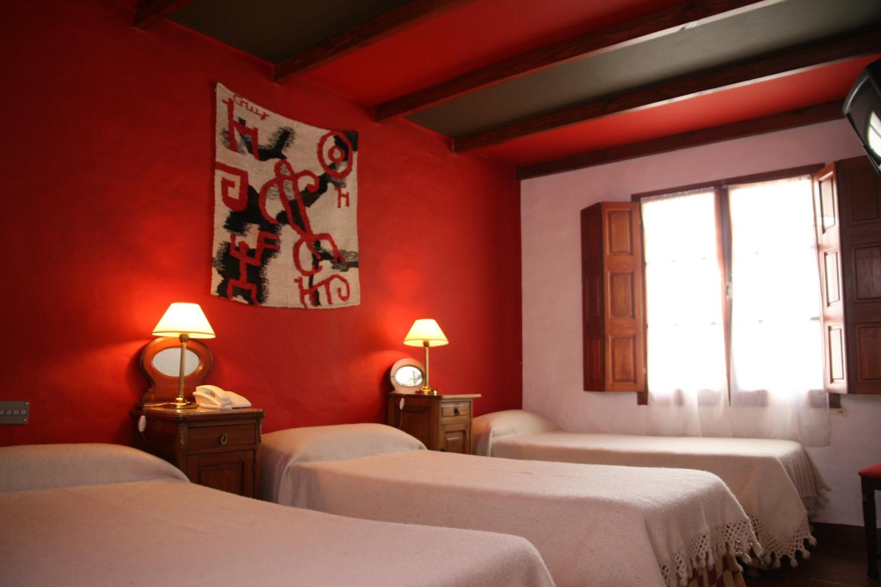 Une chambre d’hôtel avec trois lits, des meubles en bois, un mur rouge et une fenêtre avec des rideaux.