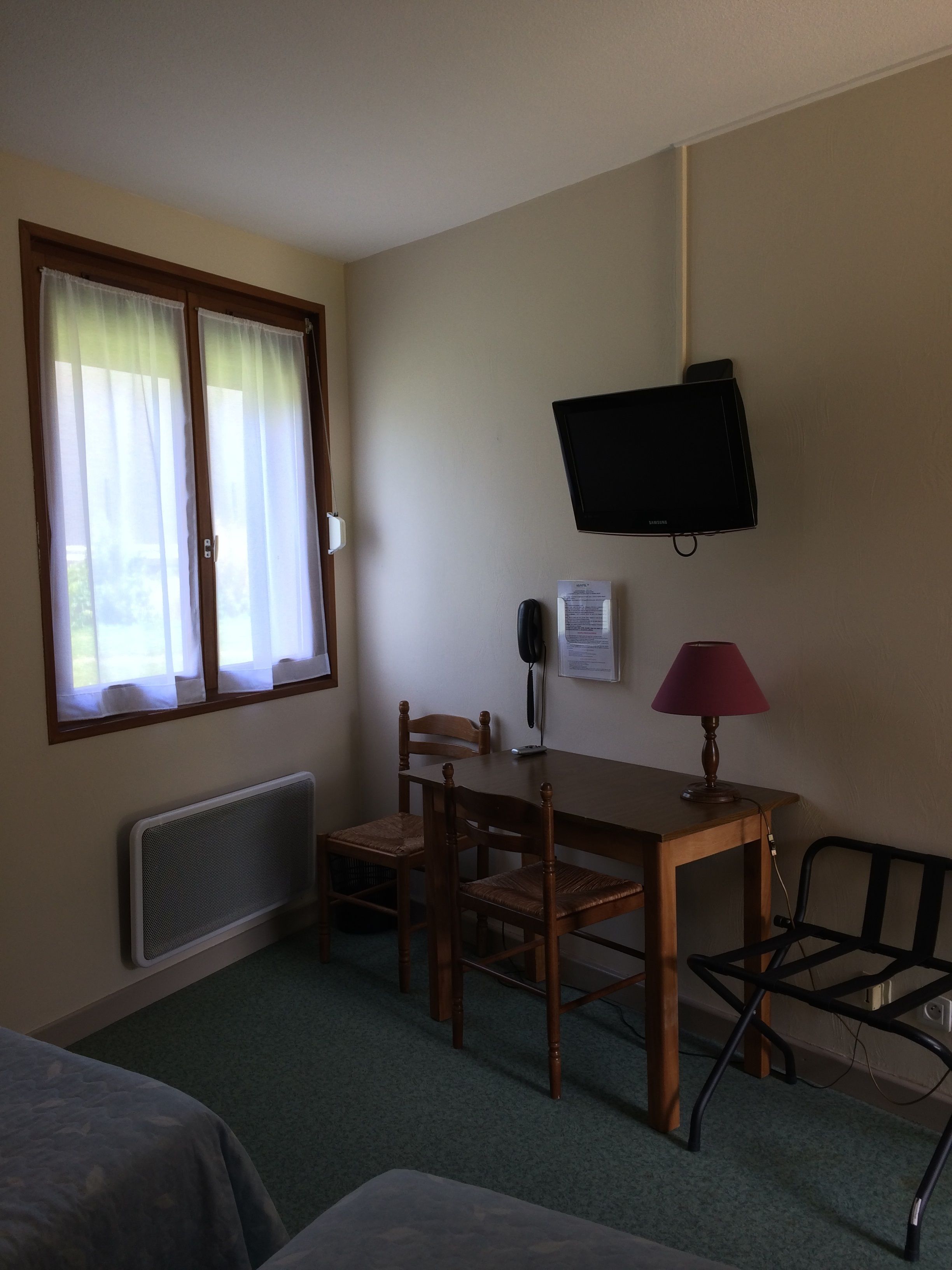 A hotel room with a wooden table, two chairs, a TV mounted on the wall, a lamp, a radiator, and a window with curtains.