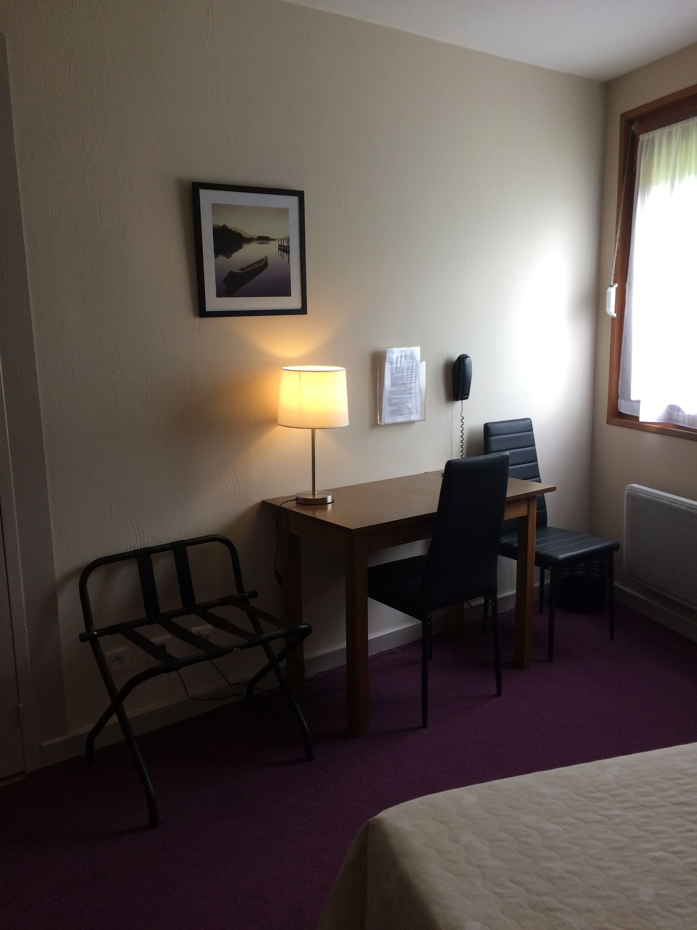 A hotel room with a bed, a desk with a chair, a lamp, a framed picture on the wall, a telephone, and a window with curtains.