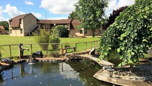 A pond with a wooden bridge, surrounded by a grassy area and a building with a sloped roof. - Delfotel (Hautefond)