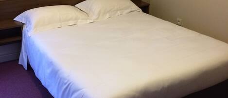 Una cama bien hecha en una habitación de hotel con cabecera de madera, alfombra morada y una lámpara fijada a la pared.