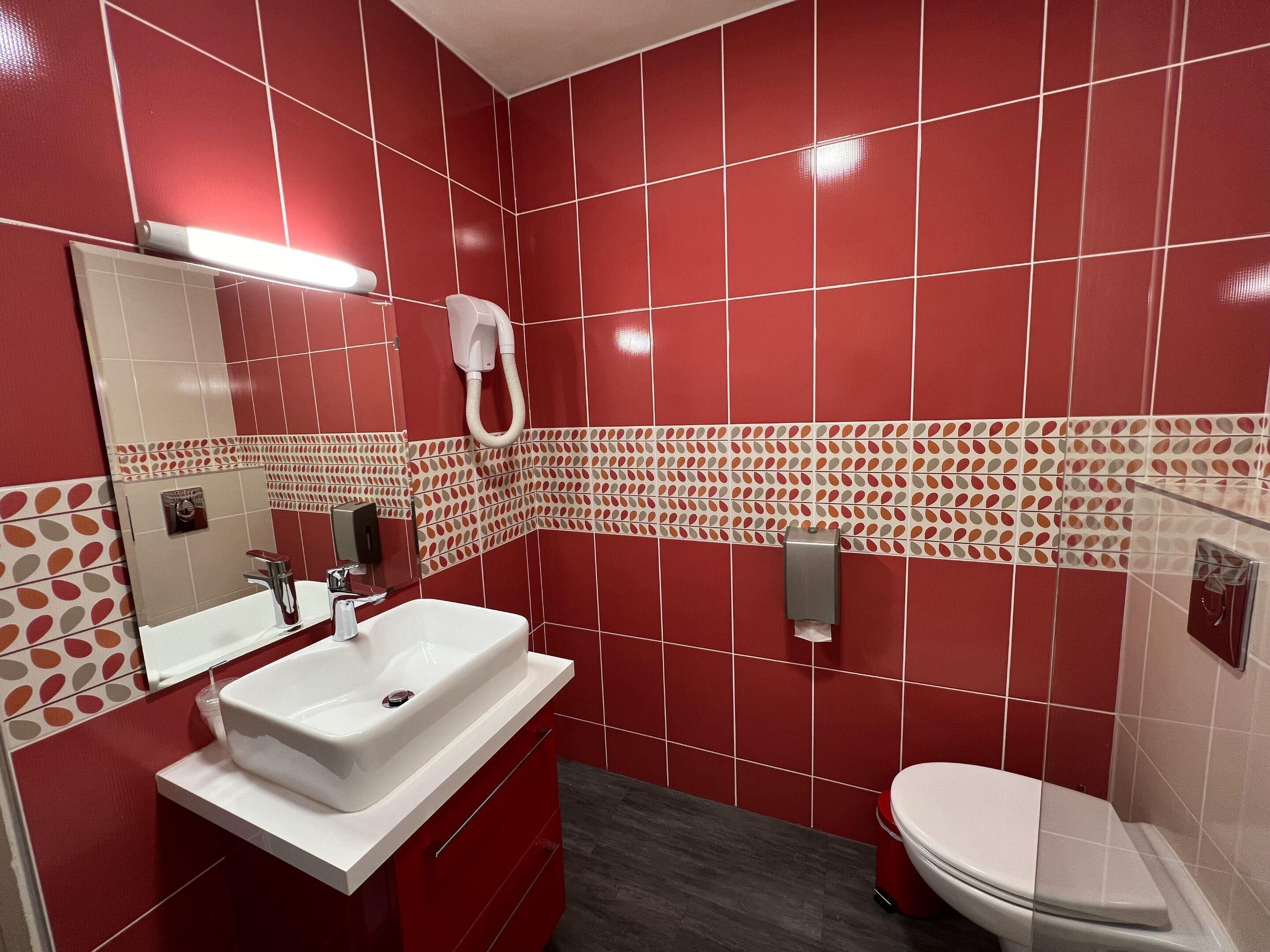 A bathroom with red tiled walls, a white sink, a mirror, a toilet, and a hand dryer.