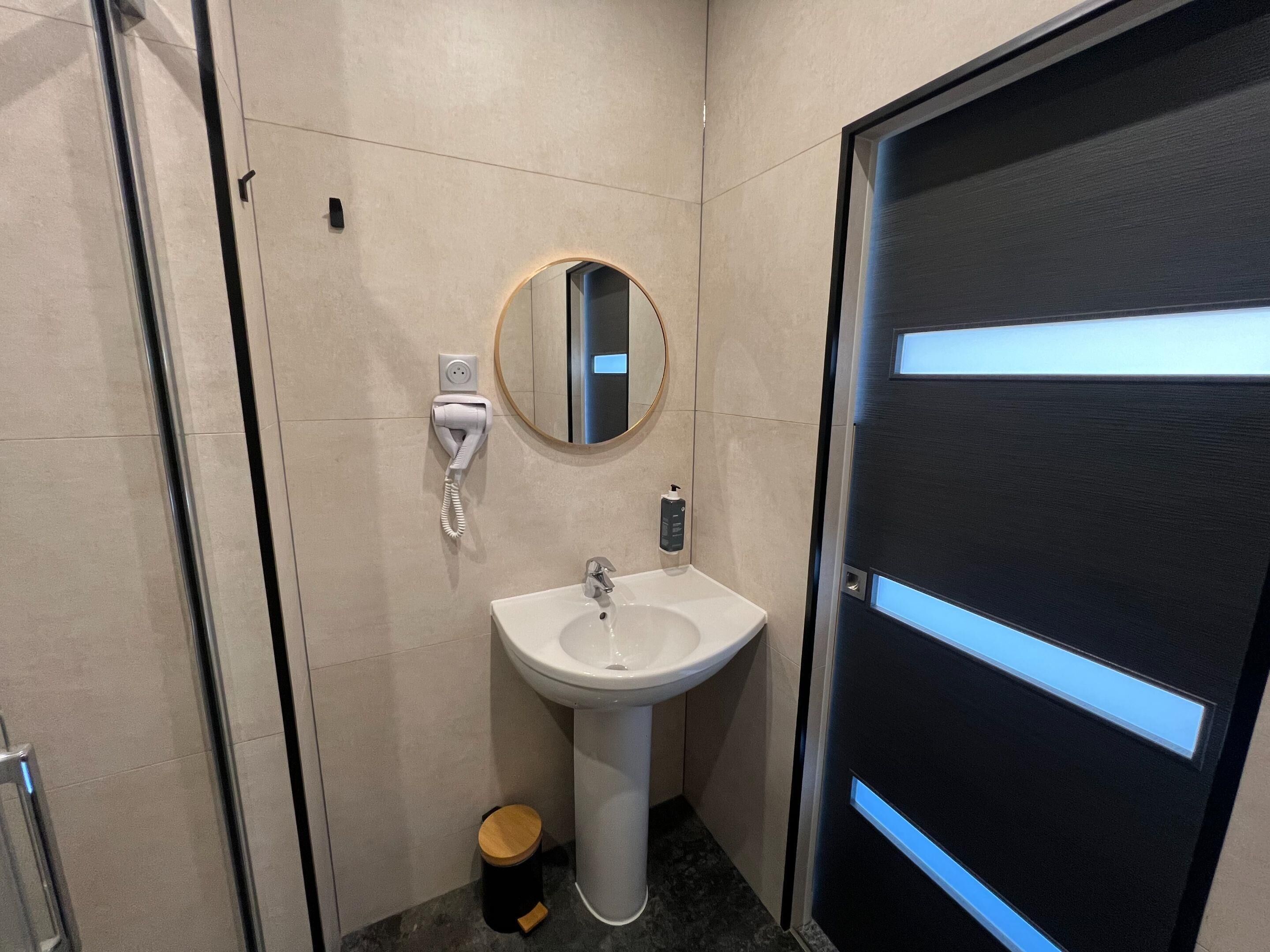 Un baño moderno con un lavamanos blanco, un espejo, un secador de manos y un cubo de basura.