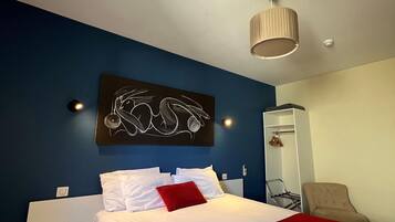 A hotel room with a bed, a red and white bedspread, a wall art piece, a ceiling light, and a chair.
