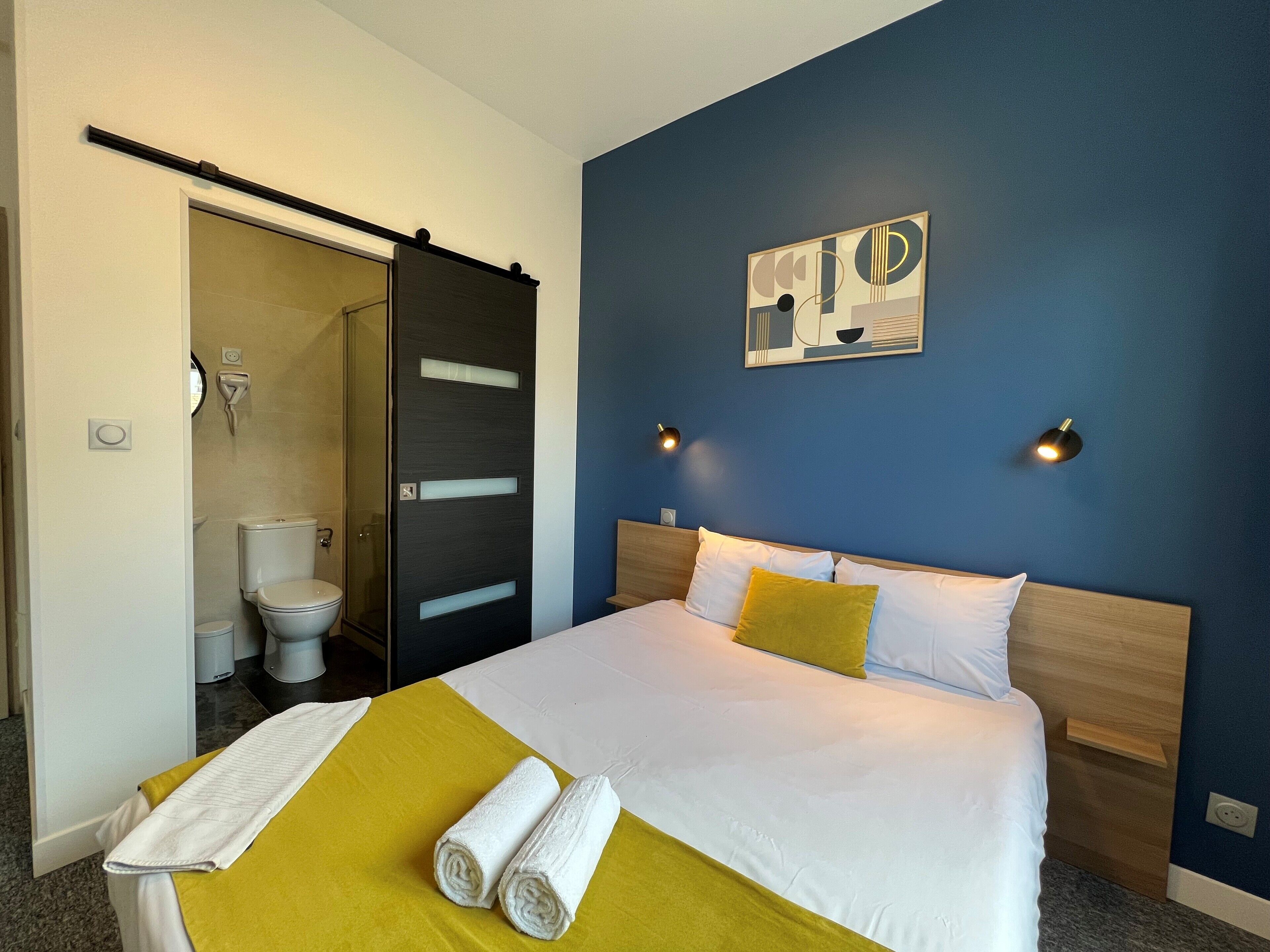 Una habitación de hotel con cama, baño y pared azul con un cuadro.
