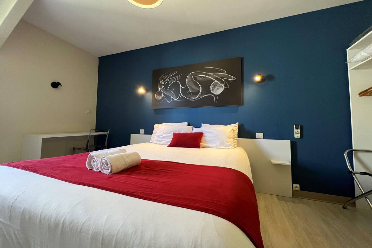 A hotel room with a large bed, a red and white bedspread, a white pillow, a red pillow, a wall-mounted artwork, and a desk with a chair.