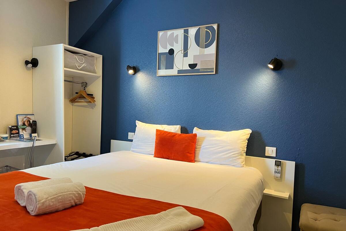 A hotel room with a bed, a desk, a wardrobe, and a wall-mounted artwork.