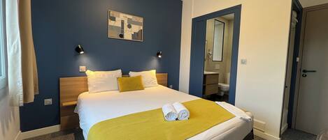 Habitación de hotel con cama, cabecero, mesita de noche, lámpara de pared, espejo, puerta y piso alfombrado.