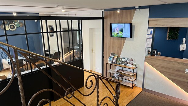A hotel reception area with a wooden reception desk, a TV mounted on the wall, and a staircase with a metal railing.