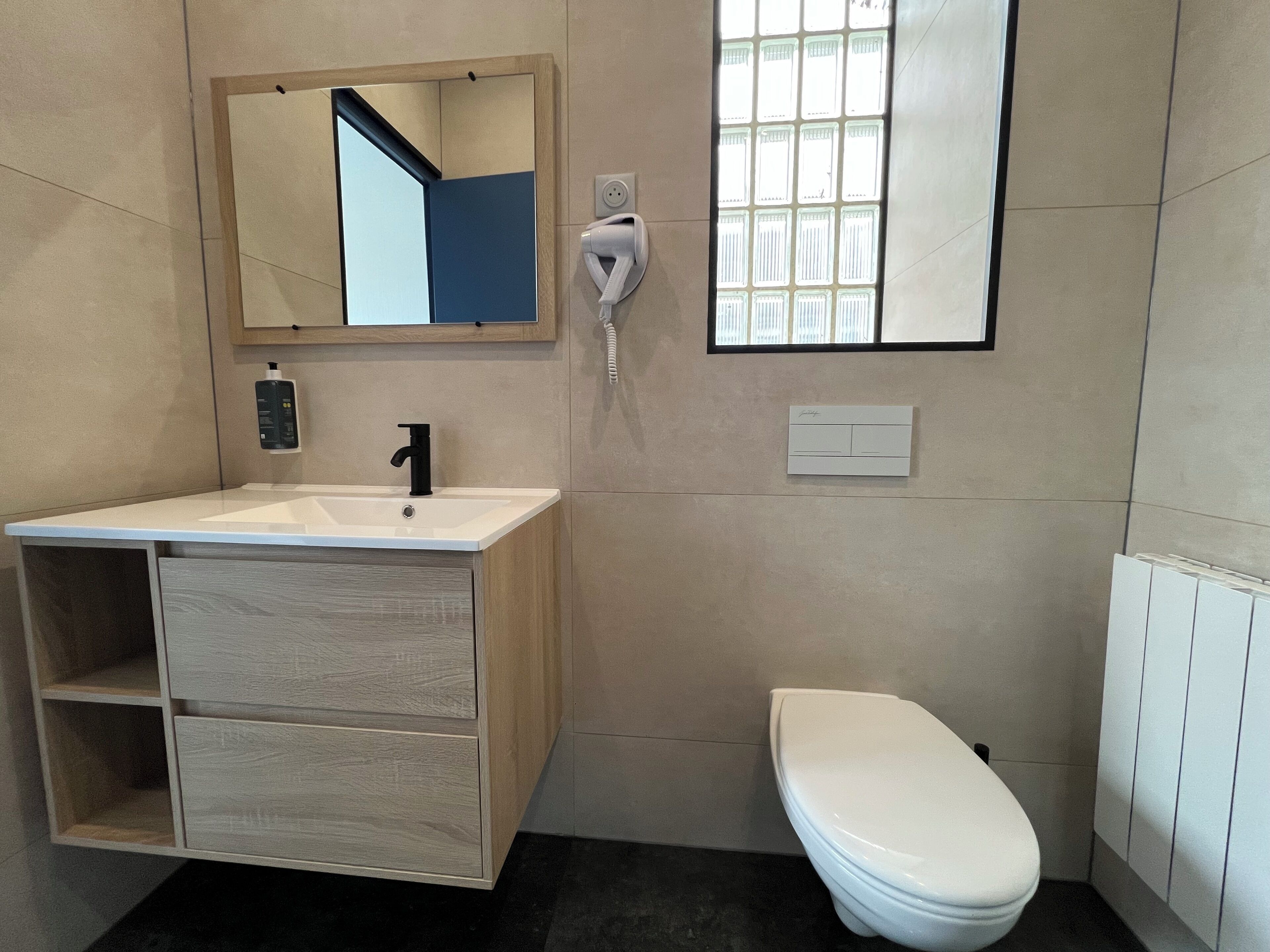 A modern bathroom with a wooden vanity, a white sink, a mirror, a hairdryer, a toilet, and a window with blinds.