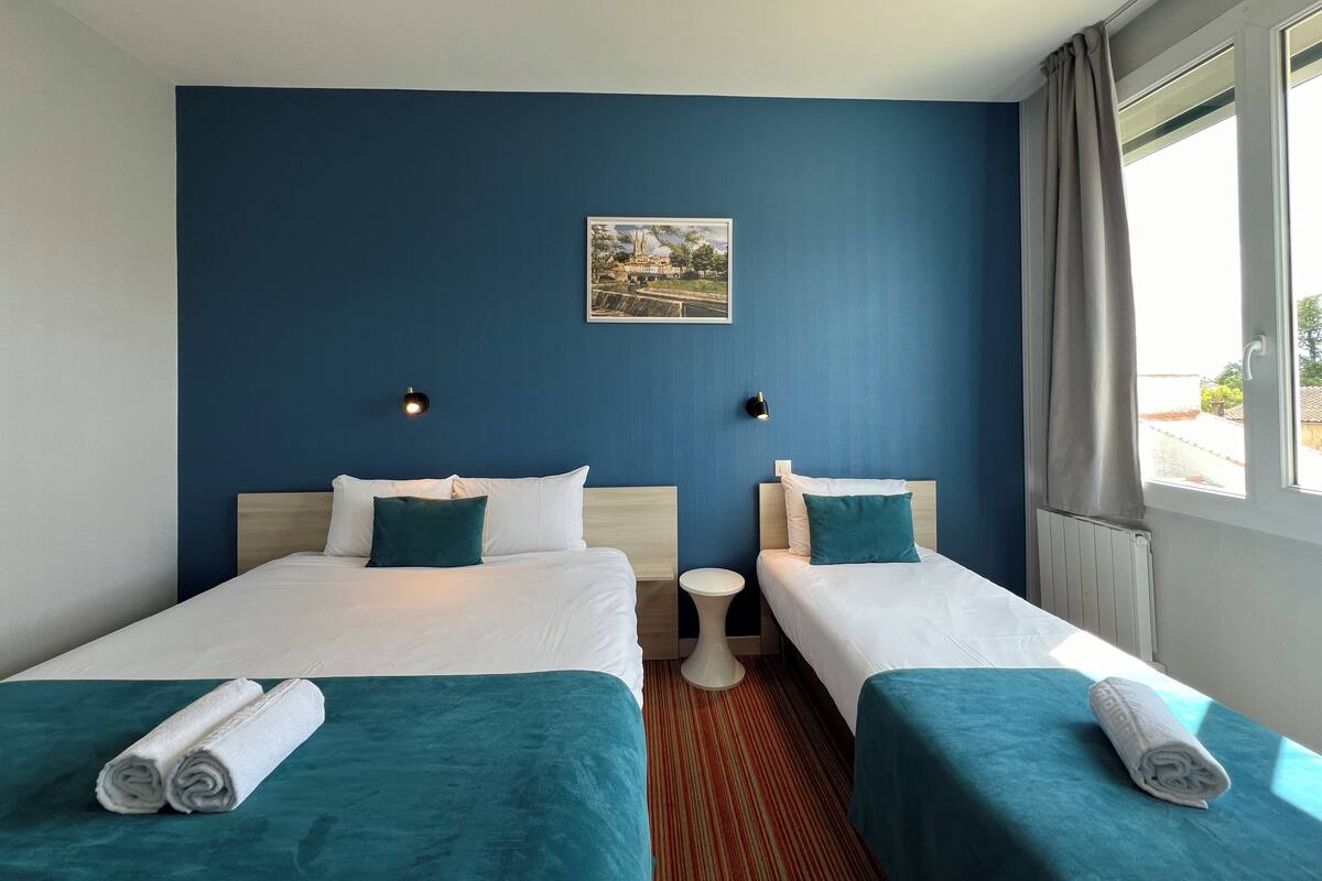 A hotel room with two beds, a small round table, a painting on the wall, and a window with curtains.