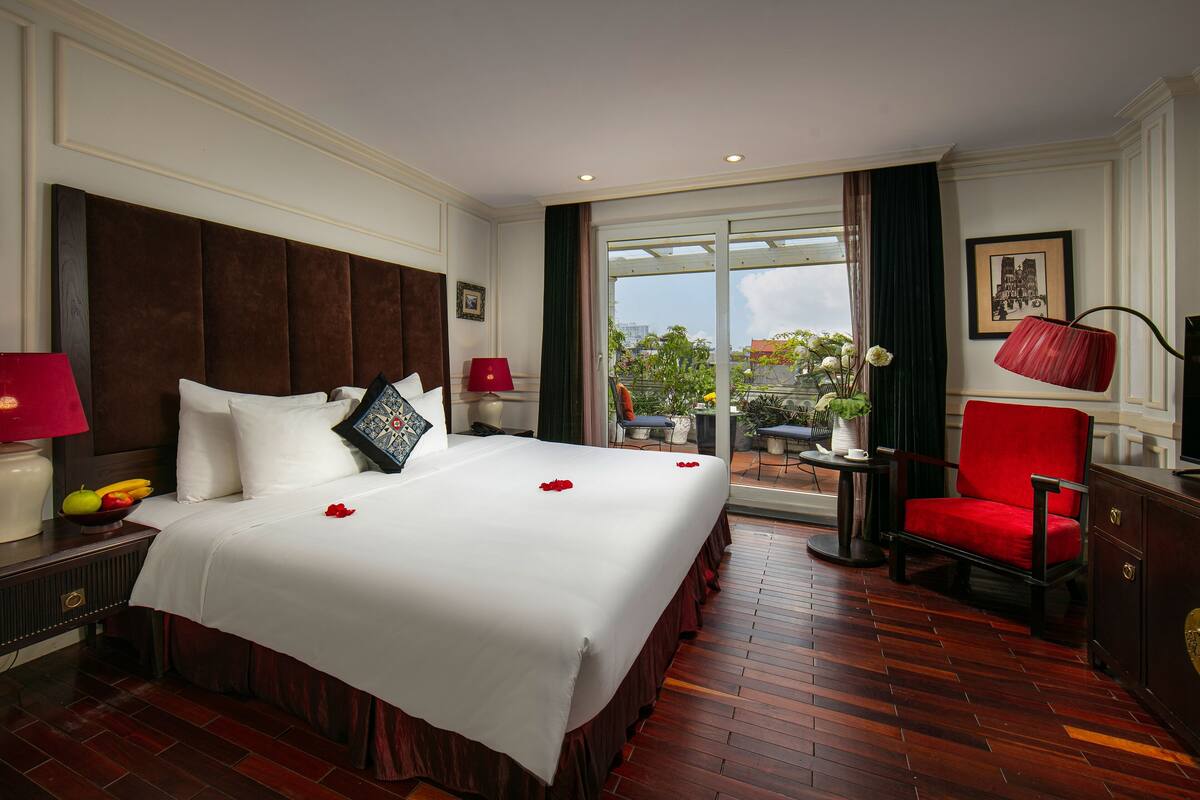 Habitación de hotel con una cama grande, una silla roja, un escritorio con lámpara y vistas al exterior.