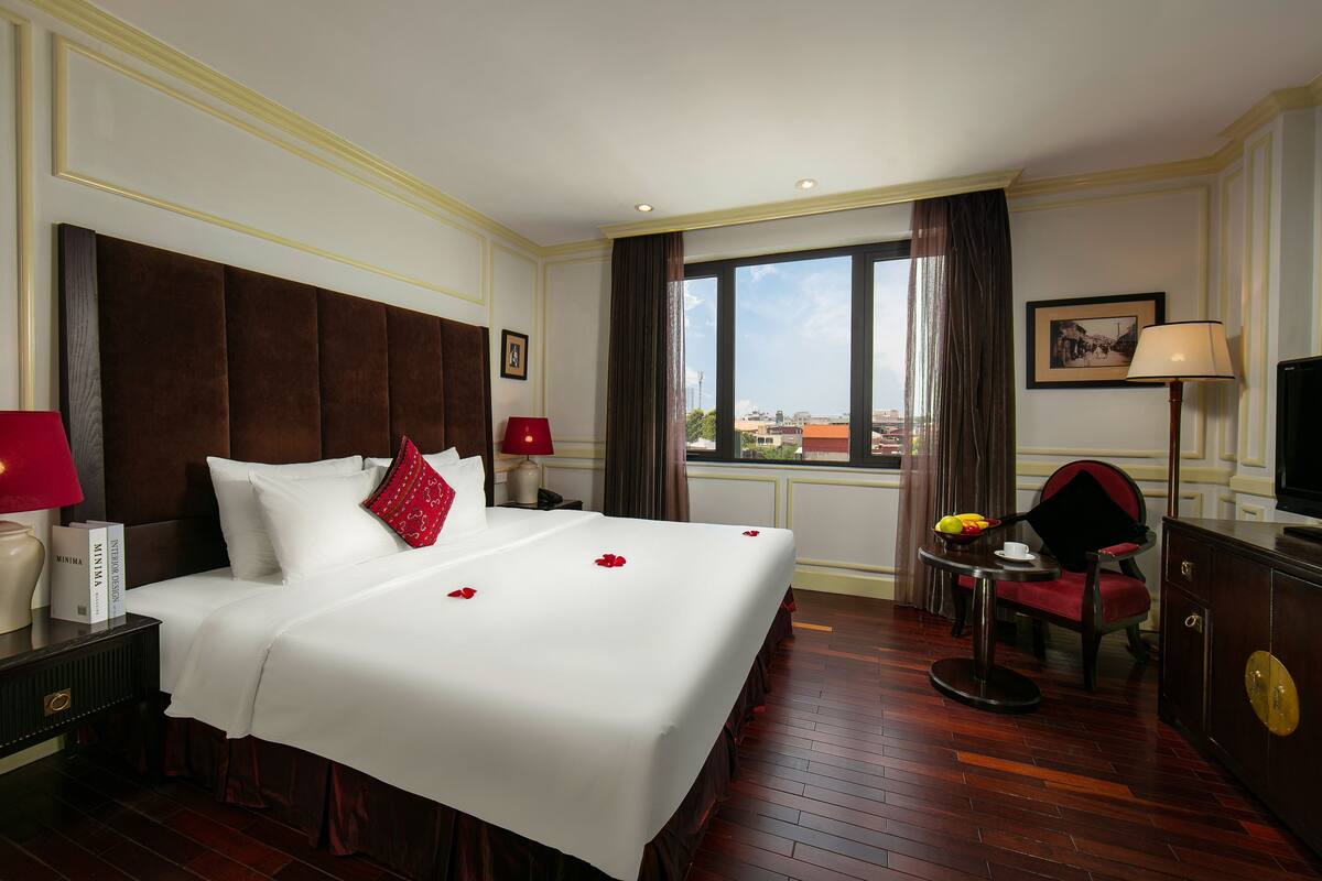 Habitación de hotel con una cama grande, dos mesitas de noche, una silla, una pequeña mesa con lámpara y un televisor.